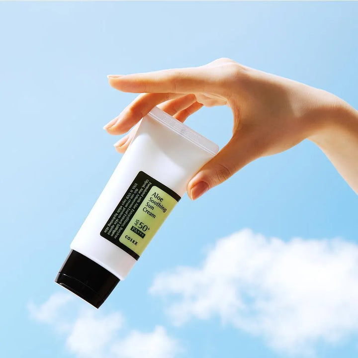 Hand holding COSRX Aloe Soothing Sun Cream SPF50 Korean skincare against blue sky