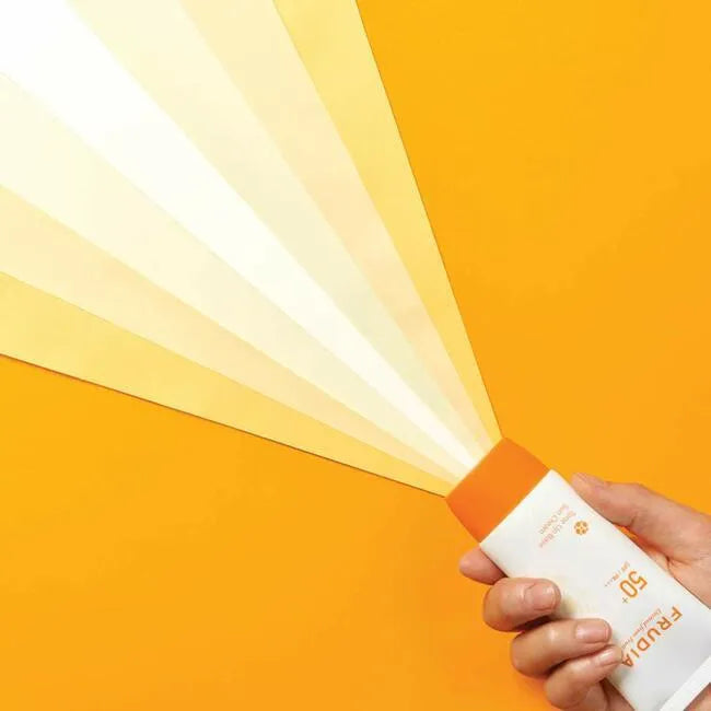 Hand holding Frudia Tone-Up Base Sun Cream SPF 50 against bright orange background, Korean skincare product at Me Time Groningen.