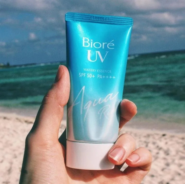 Hand holding Biore UV Aqua Rich Watery Essence SPF 50 sunscreen on a beach, Korean skincare